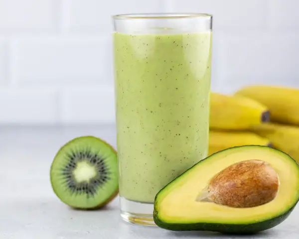 Zielony, kremowy koktajl z awokado, kiwi i banana. Obok szklanki połówka awokado i przekrojone kiwi. W tle pęk bananów.