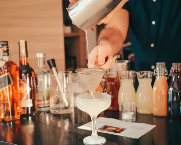 Barman przelewa płyn z shakera przez sitko do kieliszka, tworząc koktajl. W tle butelki alkoholi i kolorowe syropy.