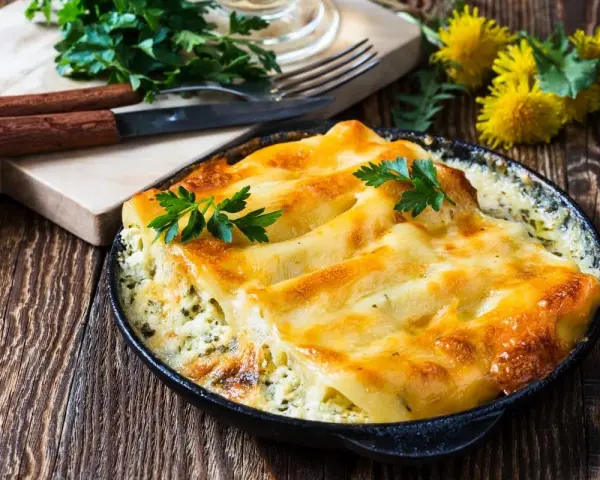 Pyszne conchiglioni zapieczone z serem i ziołami, podane w czarnej patelni. Obok deska z nożem i widelcem, oraz pęczek natki pietruszki.