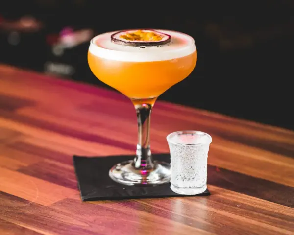 Orzeźwiający drink z marakują, ozdobiony plasterkiem owocu, stoi obok kieliszka z wodą gazowaną na drewnianym blacie.