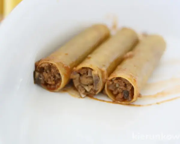 Trzy cannoloni z farszem mięsnym, wyglądające jak naleśniki z mięsem, leżą na białym talerzu.