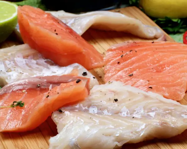 Stek z łososia kontra polędwica z dorsza. Surowe kawałki ryb, przyprawione pieprzem, leżą obok limonki, cytryny i ziół.