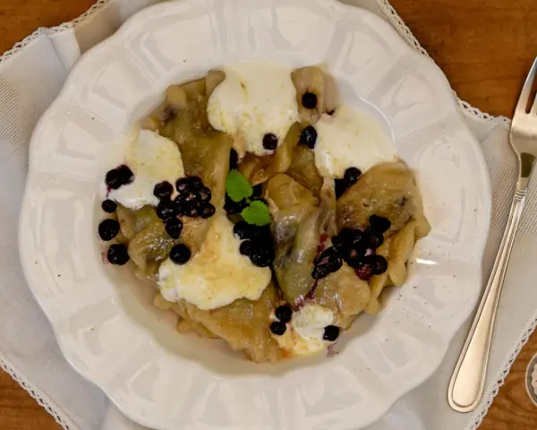 Knedle ze śliwkami czy pierogi z jagodami? Na talerzu pyszne pierogi z jagodami, polane śmietaną i posypane świeżymi owocami.