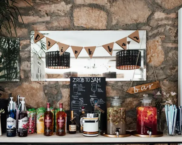 DIY drink bar z butelkami alkoholu, sokami i dzbankami z napojami. Tabliczka "ZRÓB TO SAM" zachęca do tworzenia własnych drinków.