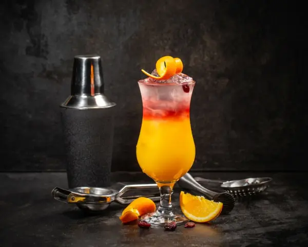 Kolorowy drink z lodem, pomarańczą i żurawiną, obok shakera. Wybór między Cosmopolitan a Sex on the Beach.