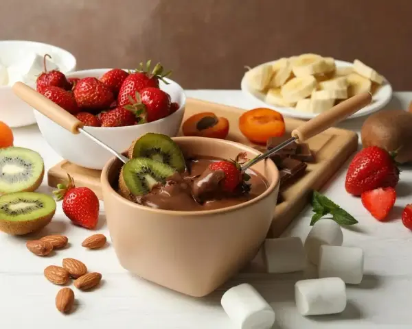 Jak zrobić fondue czekoladowe: zanurzaj truskawki i kiwi w gorącej czekoladzie. Na stole owoce, pianki i migdały.