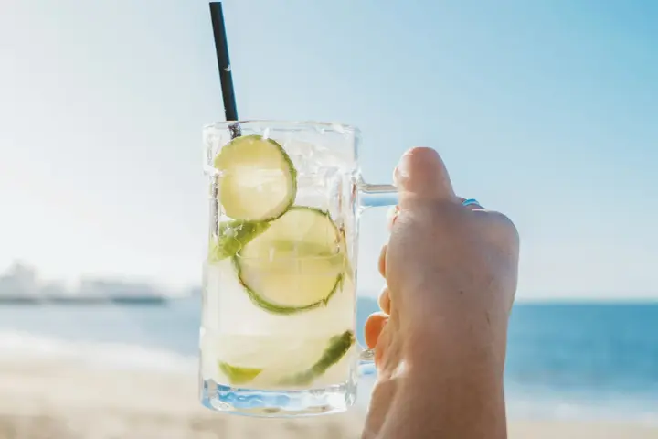 Orzeźwiająca Caipirinha czy Caipiroska z limonką i lodem, trzymana na tle plaży i błękitnego nieba. Idealny napój na wakacje.