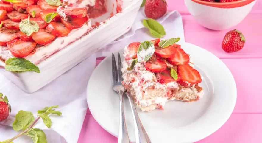 Tiramisu klasyczne kontra tiramisu truskawkowe. Porcja deseru z truskawkami i miętą na talerzu, obok naczynie z deserem i miseczka owoców.