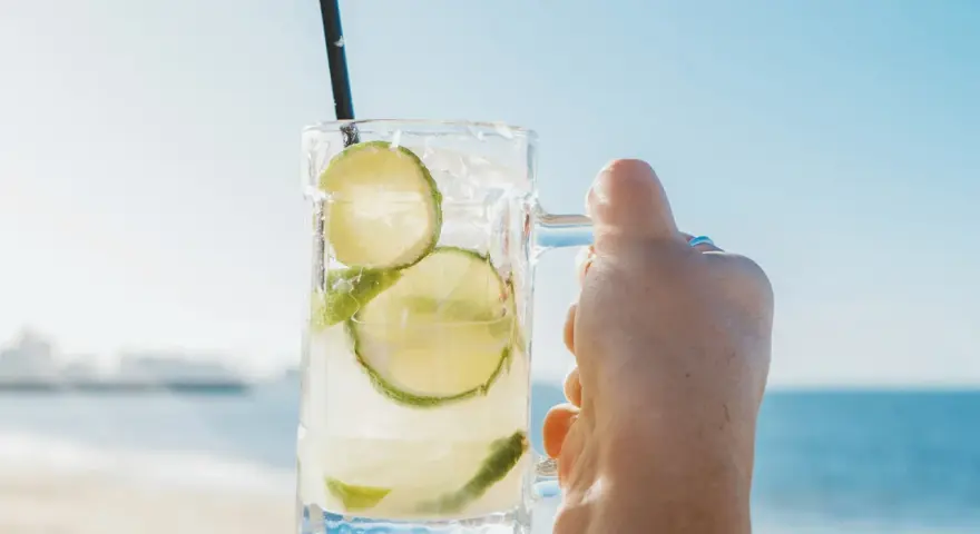 Orzeźwiająca Caipirinha czy Caipiroska z limonką i lodem, trzymana na tle plaży i błękitnego nieba. Idealny napój na wakacje.