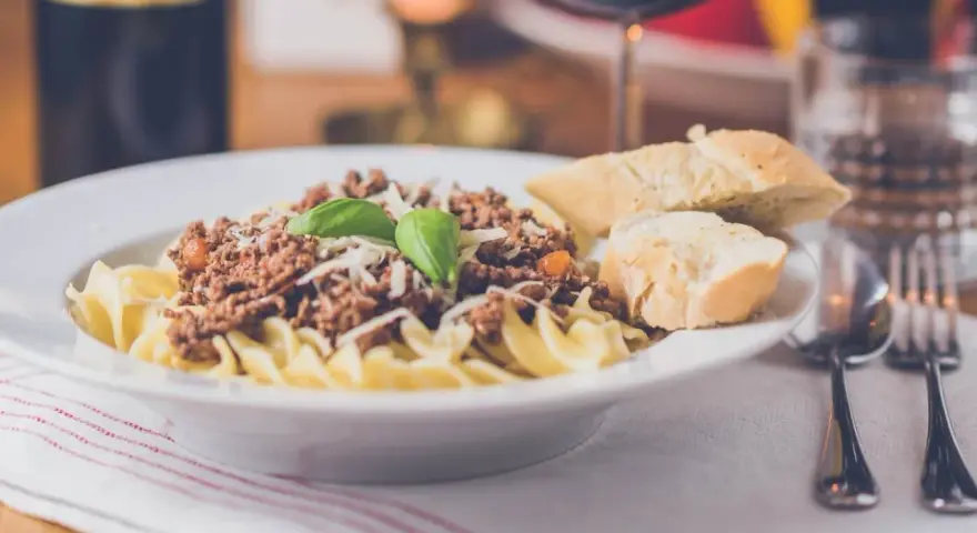 Pyszny makaron z sosem bolognese, posypany parmezanem i udekorowany bazylią. Czy to spaghetti, czy tagliatelle? Obok kromki chleba i kieliszek czerwonego wina.
