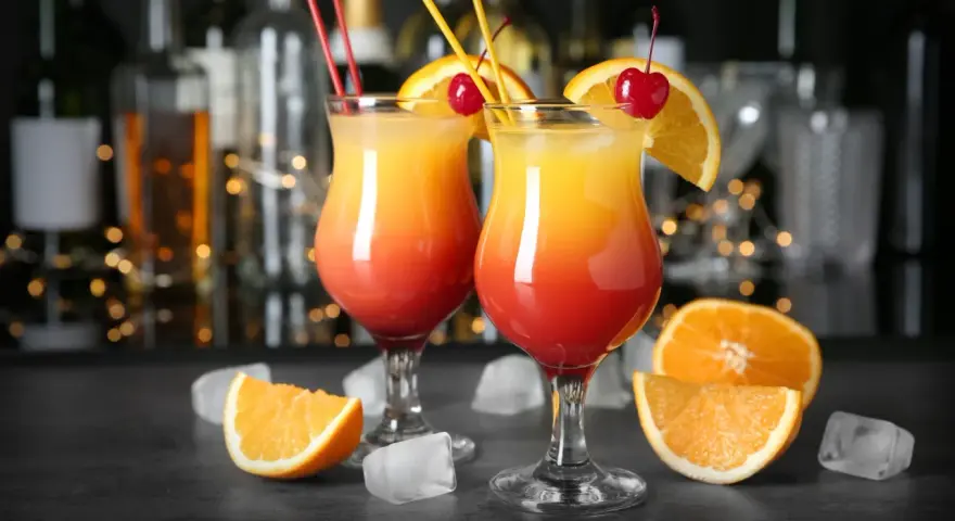 Dwa kolorowe drinki z parasolką, ozdobione pomarańczą i wiśnią, stoją na barze.