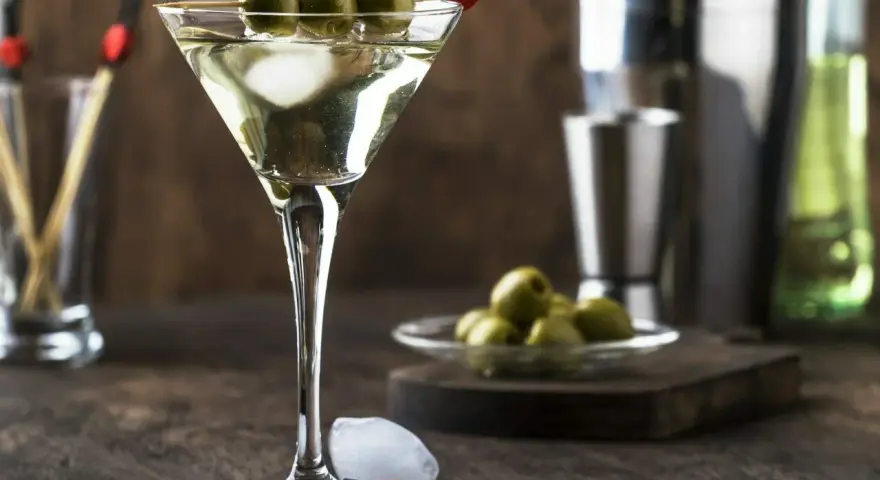 Elegancki drink z martini bianco, ozdobiony trzema oliwkami na patyczku. Obok kostki lodu i talerzyk z oliwkami.