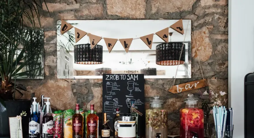 Stół z napojami i składnikami do drinków, tworzący domowy co zamiast drink baru. Na półce stoją butelki alkoholu, syropy, kartony z sokami i dwa duże słodkie poncze.