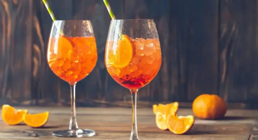 Dwa orzeźwiające drinki z aperolem, ozdobione plastrami pomarańczy i słomkami, na drewnianym blacie.
