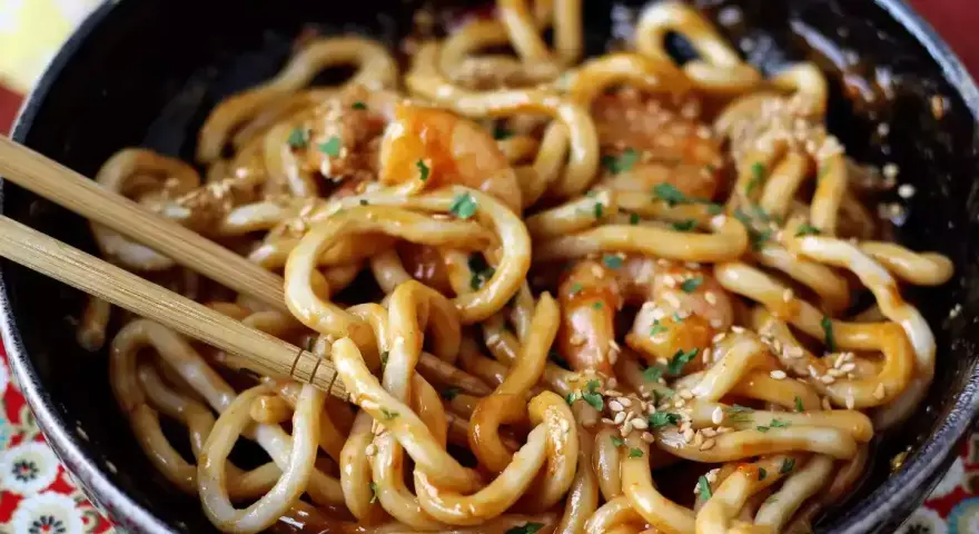 Miseczka z makaronem udon z krewetkami, posypana sezamem i pietruszką. Pałeczki zanurzone w daniu. Prosty przepis na to, jak gotować makaron udon.