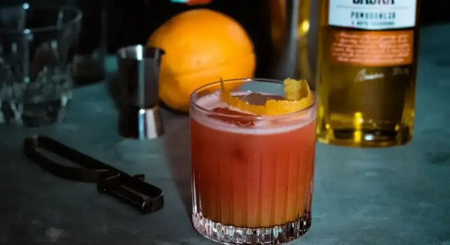 Orzeźwiający drink z likierem pomarańczowym, ozdobiony skórką cytrusową. Obok pomarańcza i butelka Saska.
