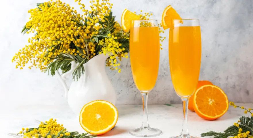 Dwa kieliszki z pomarańczowym napojem, ozdobione plasterkami pomarańczy. Obok bukiet mimozy i połówka pomarańczy. Idealne na wiosenny mimoza drink przepis.