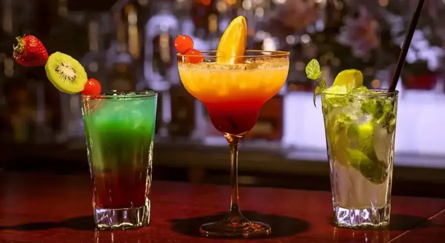 Trzy kolorowe drinki na barze. Jeden z nich to drink z pomarańczą, ozdobiony owocami.