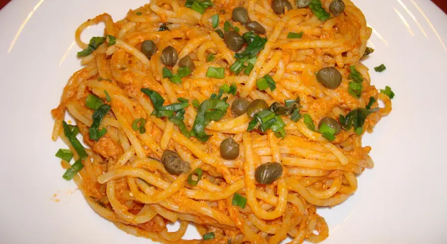 Pyszne spaghetti z tuńczykiem i kaparami, posypane szczypiorkiem, na białym talerzu.