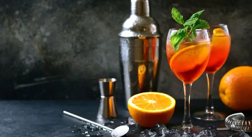 Orzeźwiający aperol z szampanem, udekorowany pomarańczą i miętą, gotowy do podania. Obok shaker i akcesoria barmańskie.