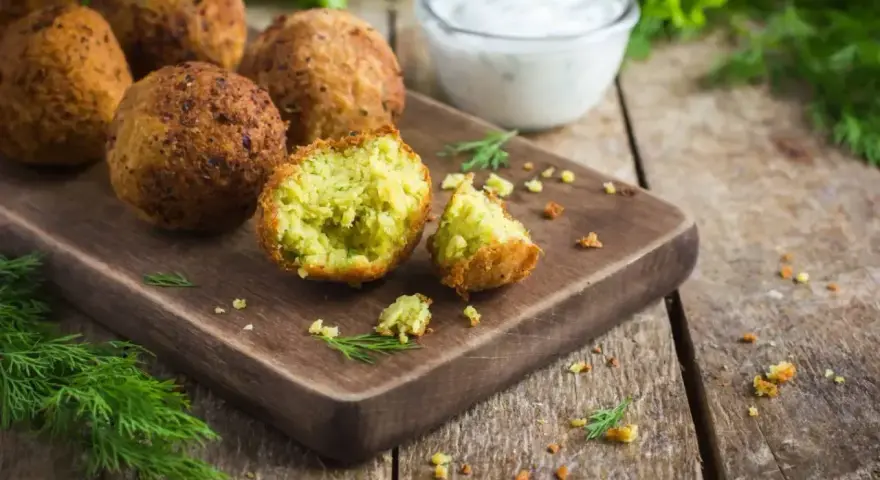 Chrupiące falafelki na desce, jeden przekrojony, ukazujący zielone wnętrze. Obok miseczka sosu i świeży koperek. Idealny falafel przepis.