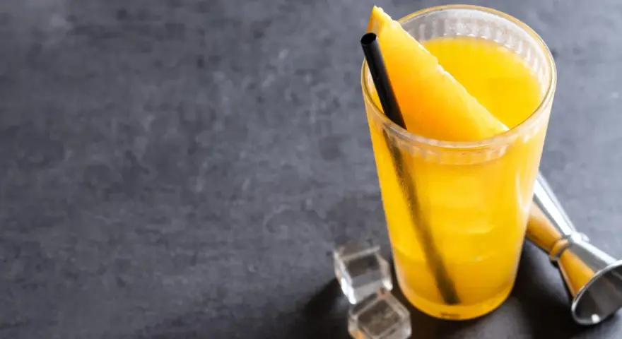 Orzeźwiający drink z wódką i sokiem pomarańczowym, ozdobiony plasterkiem pomarańczy, z kostkami lodu i miarką barmańską obok.