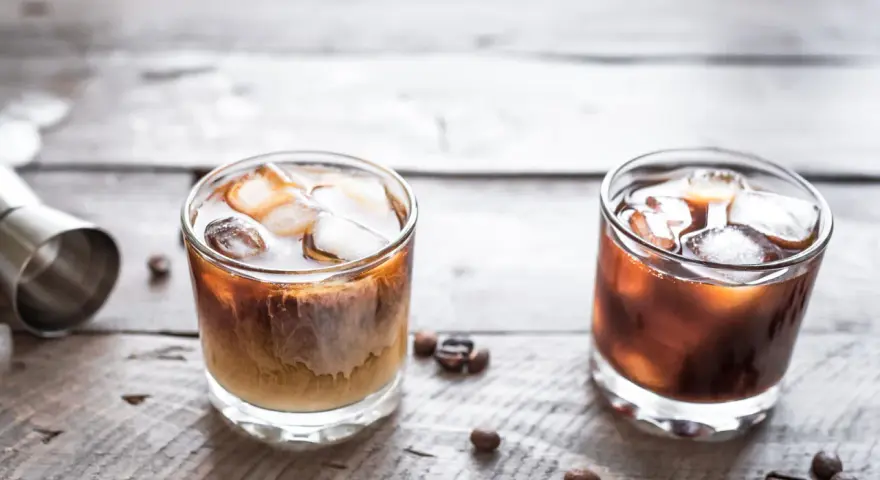 Dwa drinki na drewnianym blacie: jeden to White Russian, drugi to Black Russian.