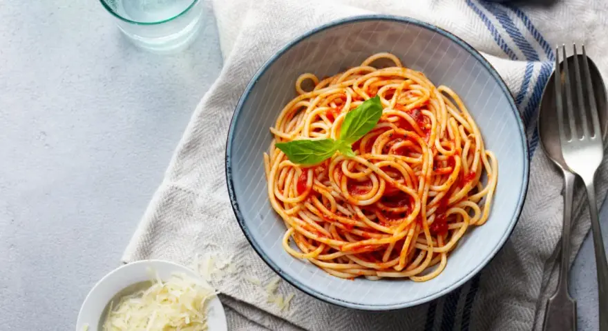 Miseczka spaghetti nr 5 z sosem pomidorowym i listkiem bazylii, obok spaghettini, starty parmezan i szklanka wody.