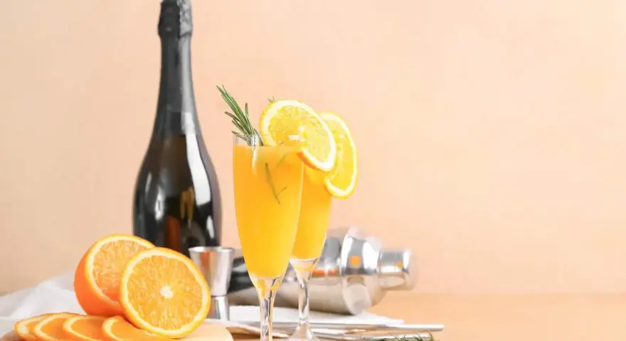 Dwa kieliszki z pomarańczowym napojem, ozdobione plasterkami pomarańczy i rozmarynem. Obok butelka szampana, shaker i świeże pomarańcze. Mimosa czy Bellini?