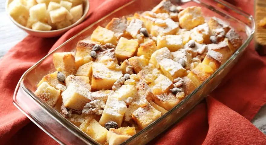 Pudding chlebowy w naczyniu, posypany cukrem pudrem i rodzynkami, obok miseczka z pokrojonym chlebem.