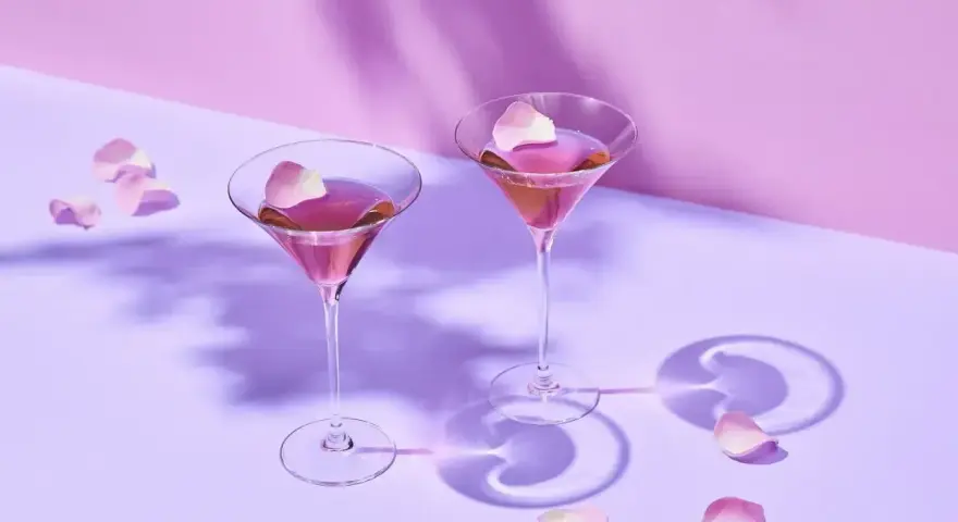 Elegancki Martini przepis: dwa kieliszki z różowym drinkiem, ozdobione płatkami róż, na fioletowym tle.