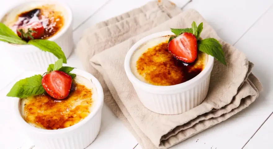 Trzy kremowe desery, Crème brûlée czy crema catalana, ozdobione truskawkami i miętą, kuszą skarmelizowanym cukrem.