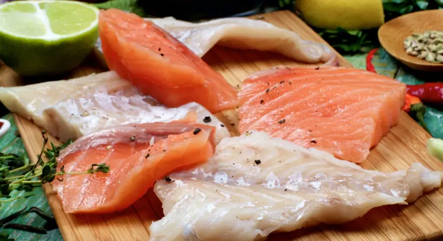 Stek z łososia kontra polędwica z dorsza. Surowe kawałki ryb, przyprawione pieprzem, leżą obok limonki, cytryny i ziół.