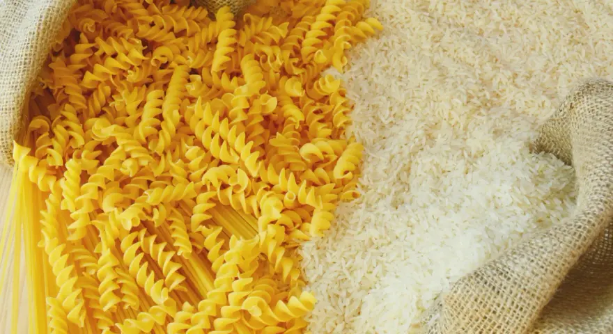 Ryż vs makaron: obok siebie leżą dwa rodzaje produktów. Z worka wysypano biały ryż, a obok niego spiralne fusilli i długie spaghetti.