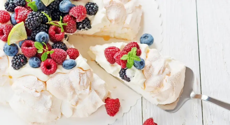 Beza Pavlova przepis: puszysta beza z bitą śmietaną i świeżymi owocami leśnymi, ozdobiona listkami mięty i plasterkami limonki.