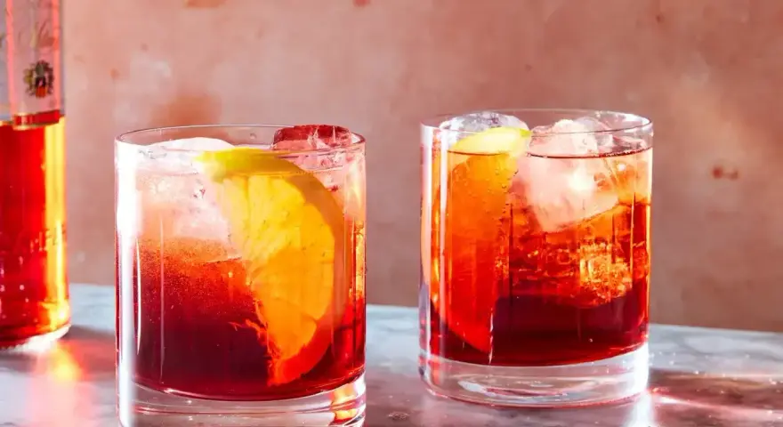 Dwa drinki Negroni z pomarańczą i lodem, obok butelki Campari. Idealny drink negroni przepis na lato.