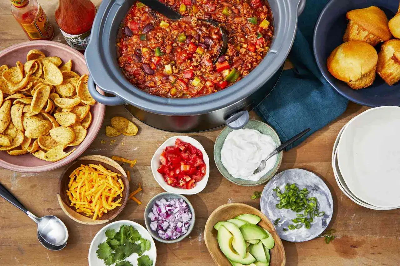 Chili con carne przepis: gorący garnek z chili, miski z nachos, serem, pomidorami, cebulą, awokado, śmietaną i szczypiorkiem.