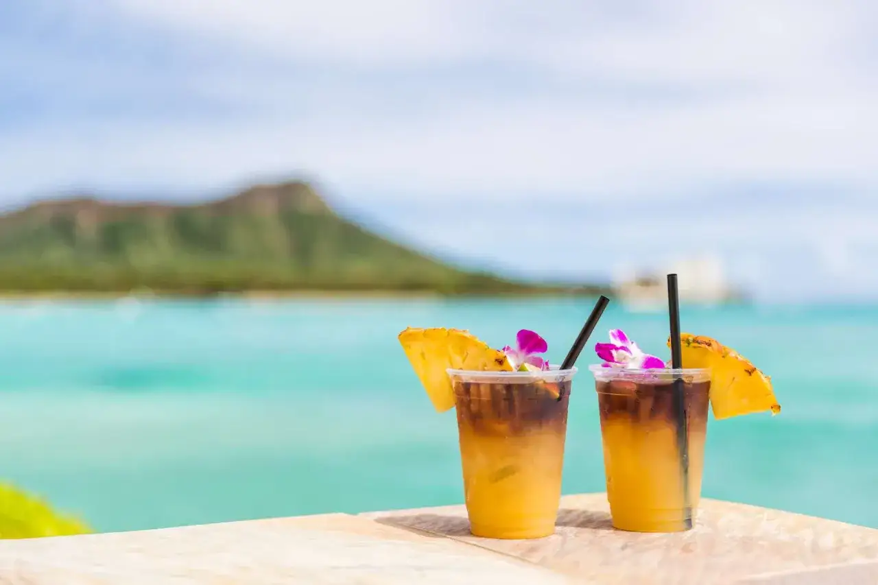 Dwa tropikalne drinki z rumem, ozdobione ananasem i kwiatami, na tle turkusowego oceanu i góry.