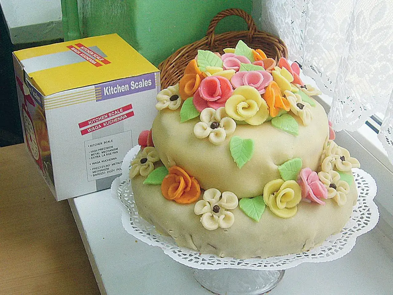 Dwupiętrowy tort ozdobiony marcepanem w kształcie kwiatów i liści. Obok stoi pudełko z wagą kuchenną.