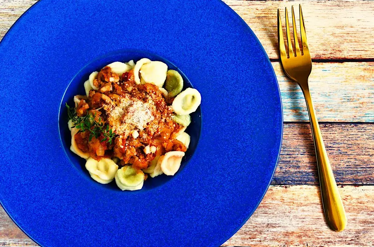 Pyszne orecchiette z sosem mięsnym, posypane parmezanem, na niebieskim talerzu. Obok złoty widelec.