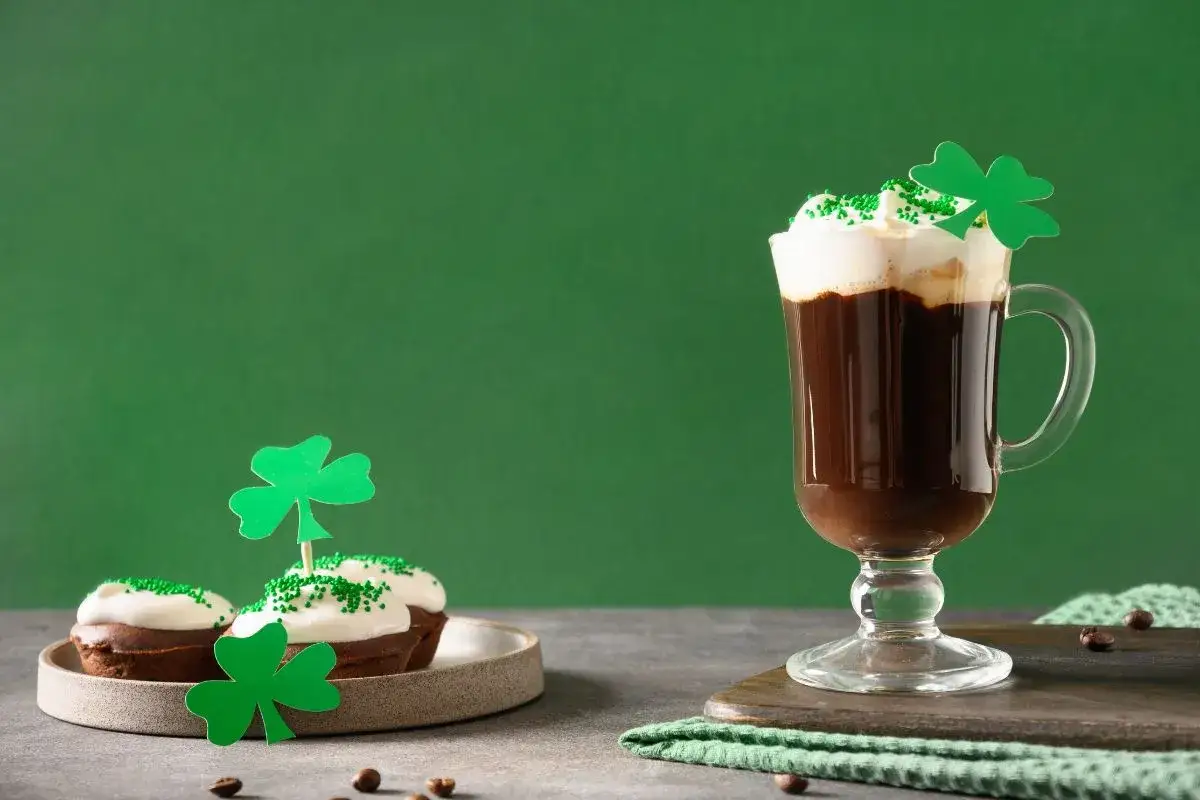 Pyszny irish coffee drink z bitą śmietaną i zielonymi posypkami, obok babeczki ozdobione koniczynkami.