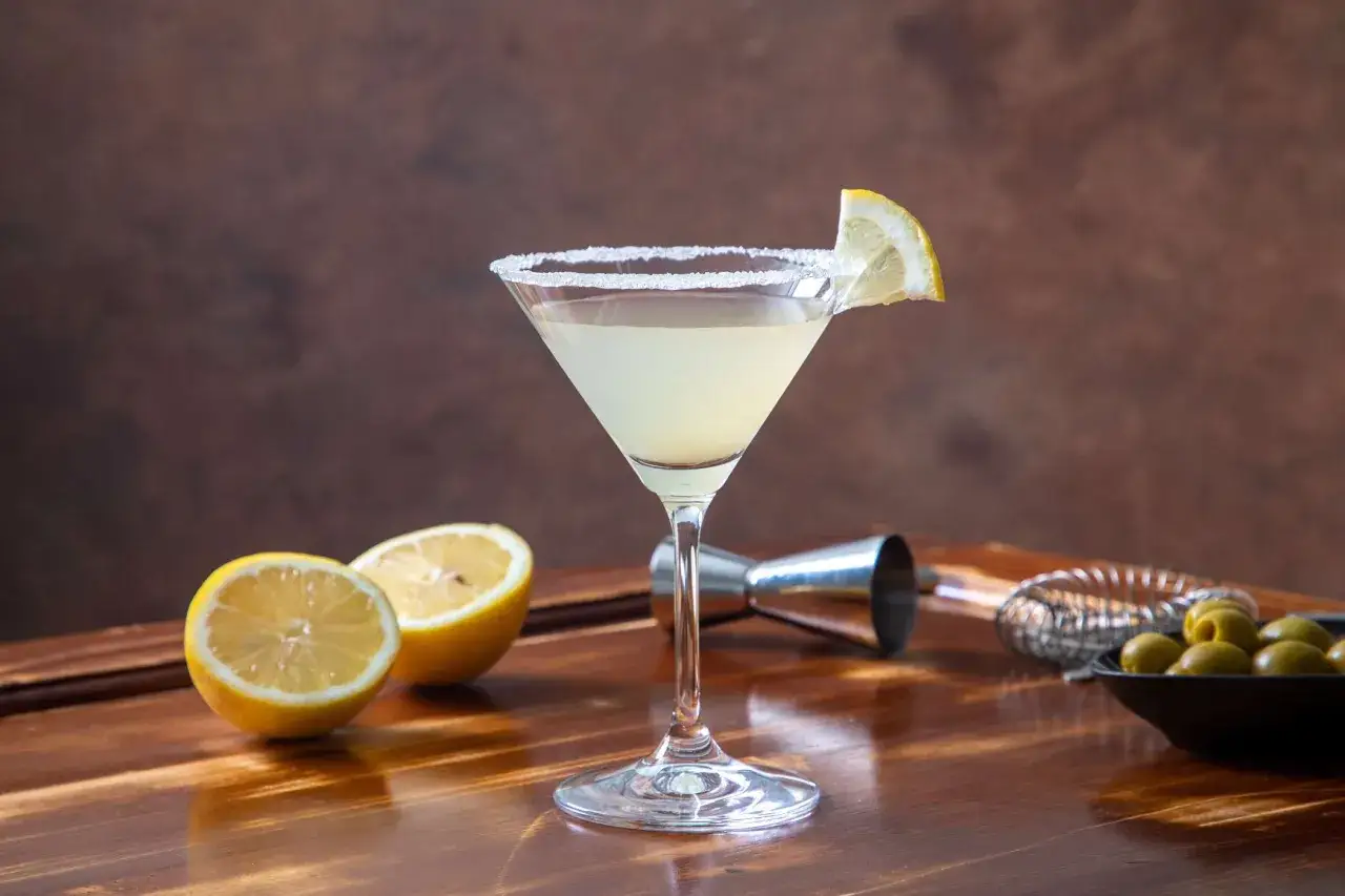 Orzeźwiający drink z martini bianco, udekorowany plasterkiem cytryny i solą na kieliszku. Obok leżą połówki cytryn i oliwki.