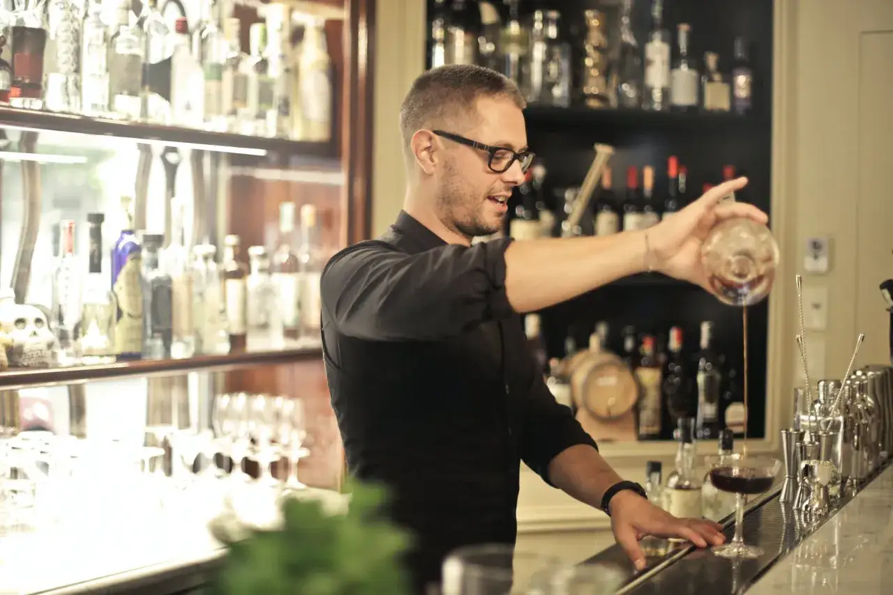 Barman w okularach nalewa drinka do kieliszka. W tle półki z butelkami alkoholu.