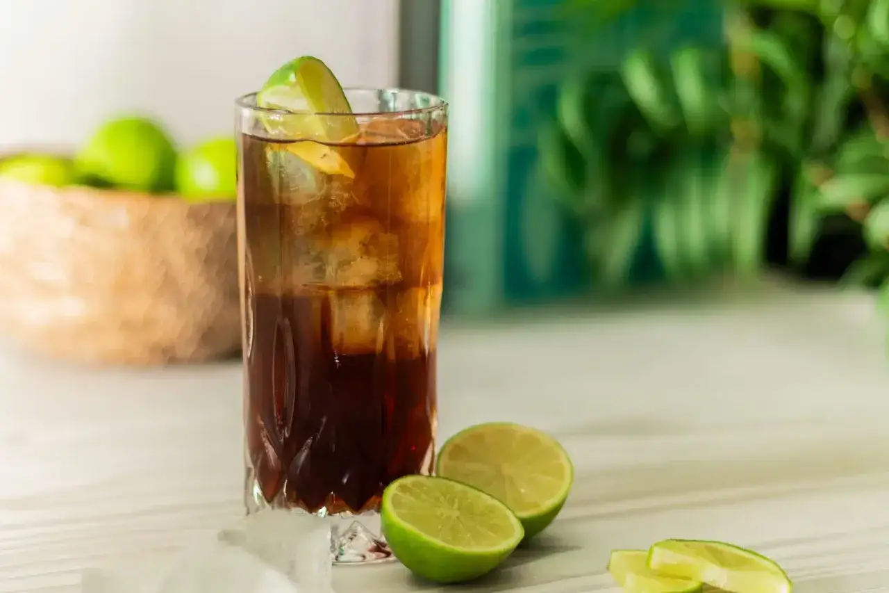 Klasyczny Cuba Libre, czyli rum z colą i limonką, w szklance z lodem. Obok świeże limonki i kostki lodu.