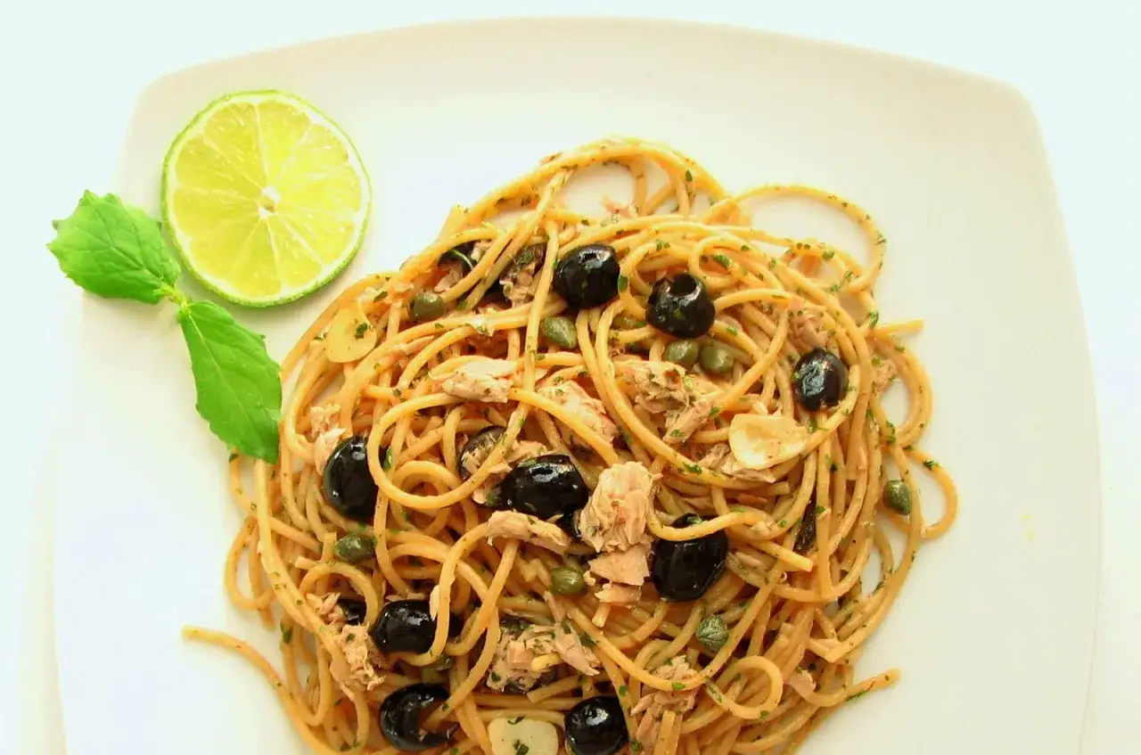 Pyszne spaghetti z tuńczykiem i kaparami, ozdobione oliwkami, czosnkiem i świeżą miętą, z plasterkiem limonki.