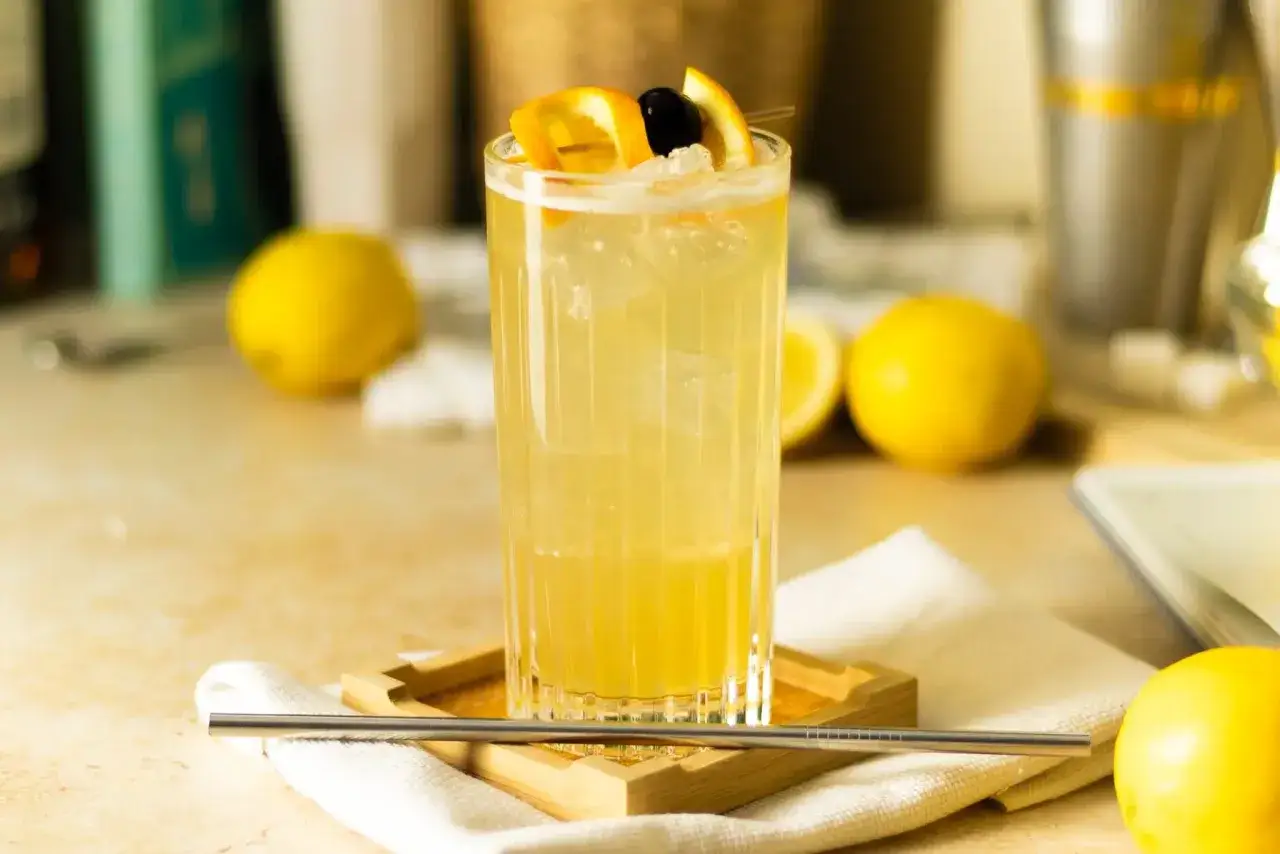 Orzeźwiający John Collins drink, ozdobiony plasterkami pomarańczy i wiśnią, otoczony cytrynami.