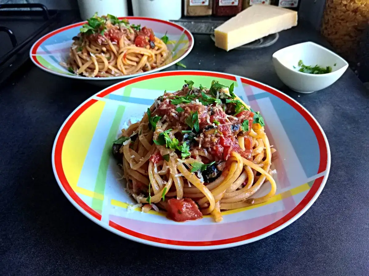 Dwa talerze z makaronem spaghetti puttanesca, posypane natką pietruszki i parmezanem. Obok kawałek sera i miseczka z ziołami.