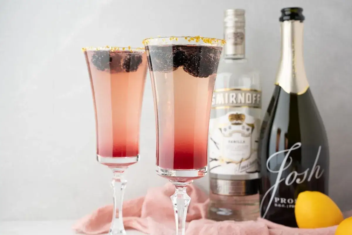 Dwa eleganckie kieliszki z różowym drinkiem z prosecco, ozdobione jeżynami i złotymi płatkami. Obok butelka wódki Smirnoff i prosecco.