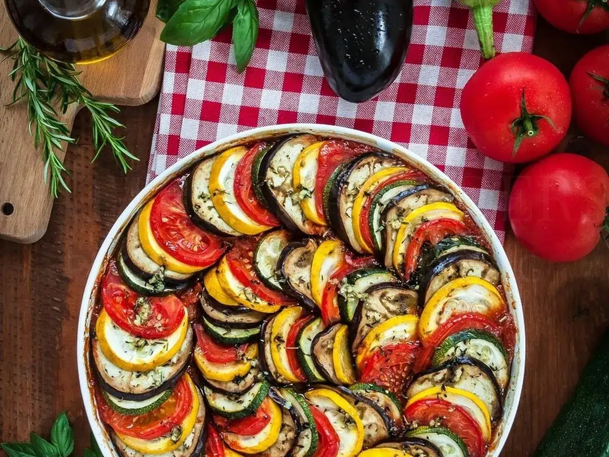 Kolorowy ratatouille przepis w naczyniu, ułożony z plastrów cukinii, bakłażana i pomidorów. Obok świeże warzywa i zioła.
