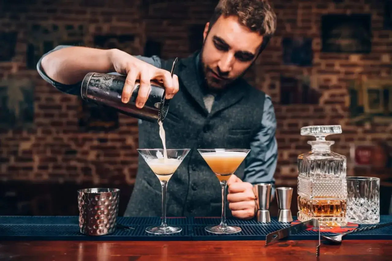 Barman przelewa drinka z shakera do kieliszka. W tle widać ceglaną ścianę i inne akcesoria barmańskie.